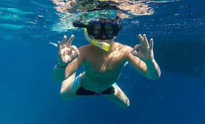 Person snorkelling in clear blue water wearing a mask and snorkel while signalling OK.